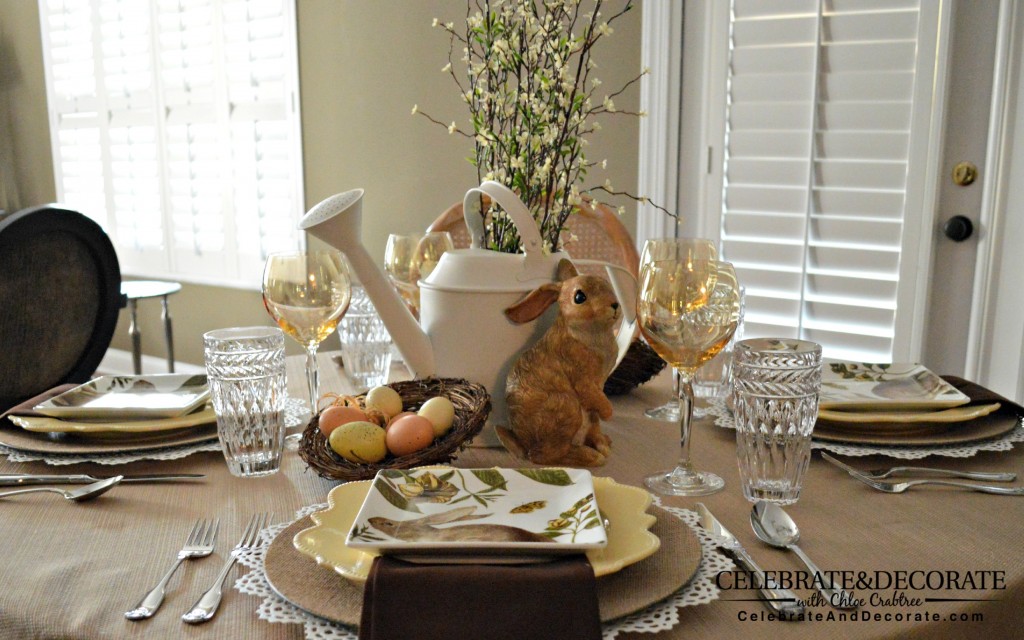 A neutral table set with white and burlap chargers, yellow plates and neutral bunny plates.  Dark brown napkins, silver flatware, yellow wine glasses, a centerpiece of a brown bunny figurine, a nest with faux eggs, a white watering can with faux blooming spring branches in it