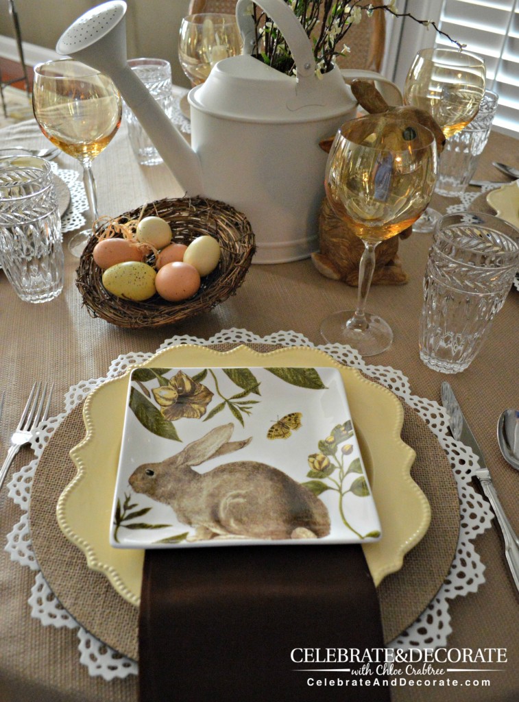 A neutral table set with white and burlap chargers, yellow plates and neutral bunny plates.  Dark brown napkins, silver flatware, yellow wine glasses, a centerpiece of a brown bunny figurine, a nest with faux eggs, a white watering can with faux blooming spring branches in it