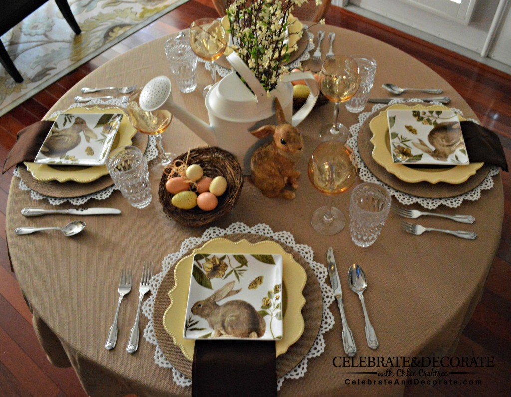 An overhead shot of A neutral table set with white and burlap chargers, yellow plates and neutral bunny plates.  Dark brown napkins, silver flatware, yellow wine glasses, a centerpiece of a brown bunny figurine, a nest with faux eggs, a white watering can with faux blooming spring branches in it