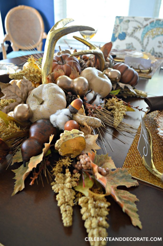 A Fall Tablescape centerpiece for Thanksgiving dinner featuring golden antlers, cotton bolls, acorns, leaves and gourds in shades of gold, cream and brown. 