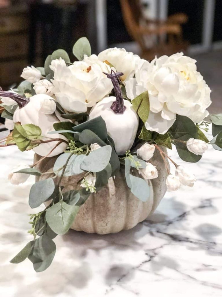 Rustic colored pumpkin with a white flower and eucalyptus floral arrangement in the pumpkin and a couple of white faux pumpkins in the arrangement 