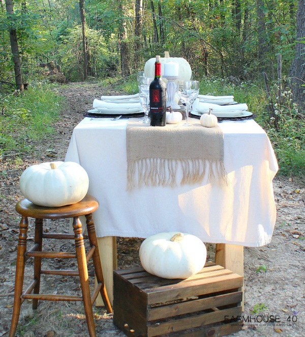 beautiful table set in the out of doors with a crisp white tablecloth, a burlap table runner, adorned with white pumpkins and a bottle of red wine