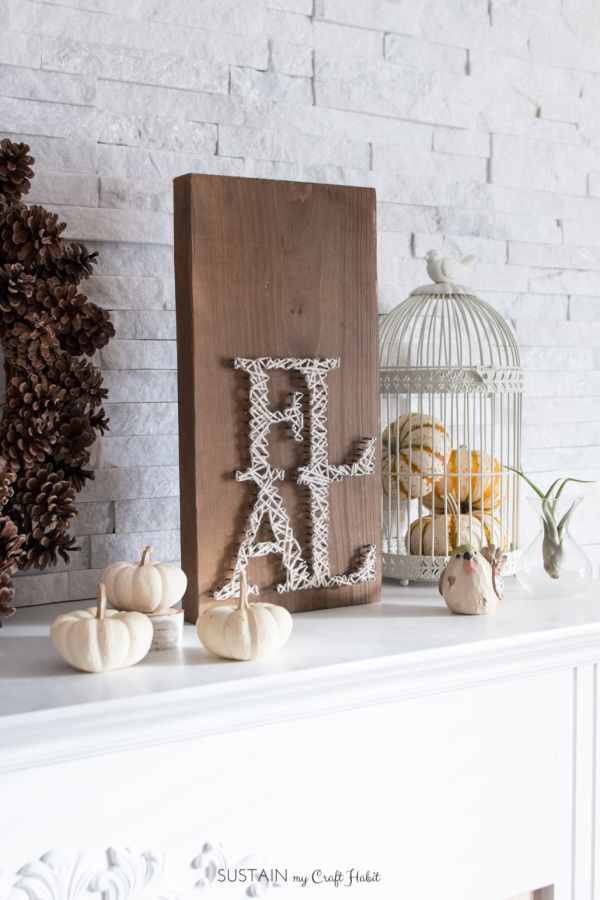 neutral fall vignette with white pumpkins, a white birdcage filled with pumpkins a white brick wall a pinecone wreath and a FALL string art craft project