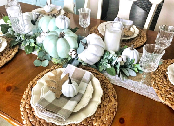 pretty wood dining table set with seagrass placemats and topped with Pioneer woman ivory dinner plates and topped with a neutral buffalo plaid napkin and white pumpkin and a paper oak leaf with a centerpiece of green and white pumpkins, white candles and greenery