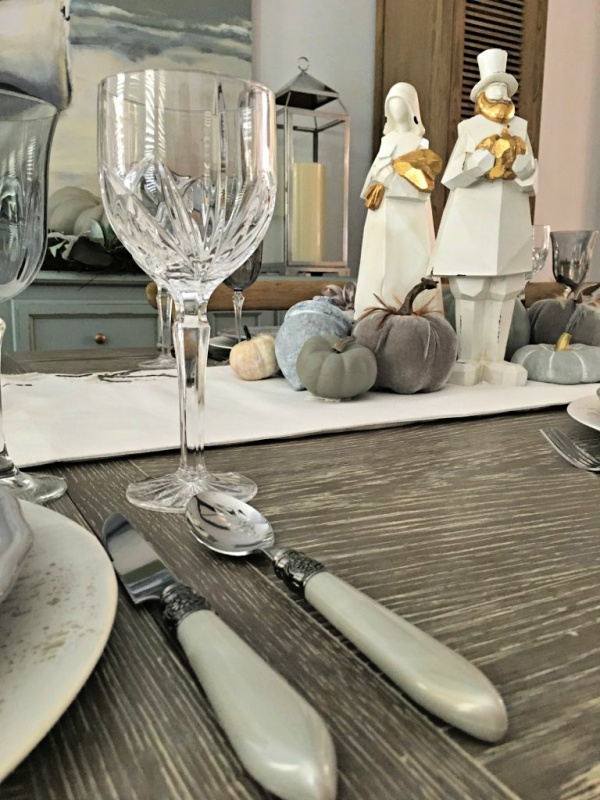thanksgiving tablescape with gray flatware gray pumpkins, waterford goblets and a pair of modern white pilgrims with gold accents