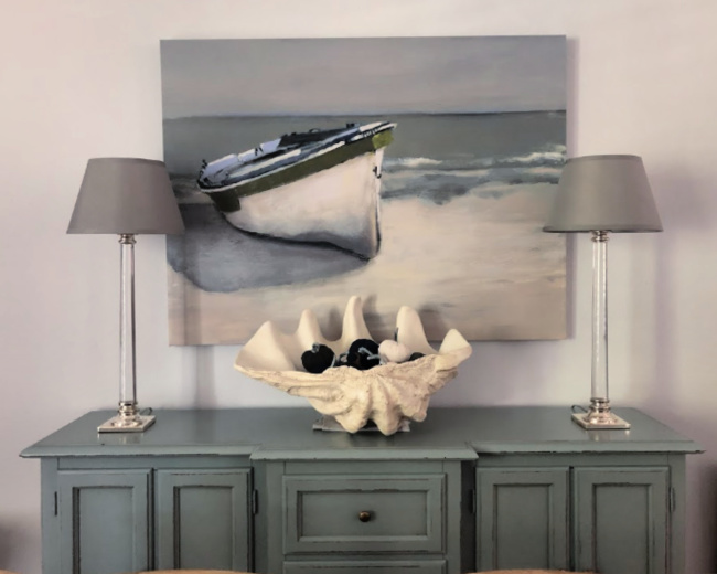 Blue sideboard with a painting of a boat above it, candlestick lamps on it and a giant clamshell filled with blue and gray pumpkins