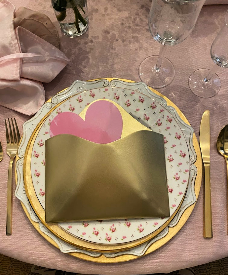 A place setting for Valentine's day with a gold envelope on the plates with a pink heart note in it
