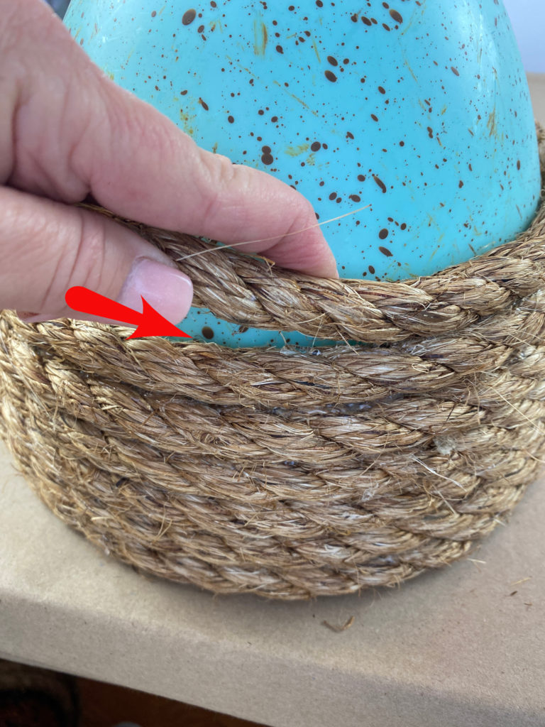 A blue decorative easter egg with jute rope being glued around it.  a red arrow and a hand wrapping the rope around the egg.