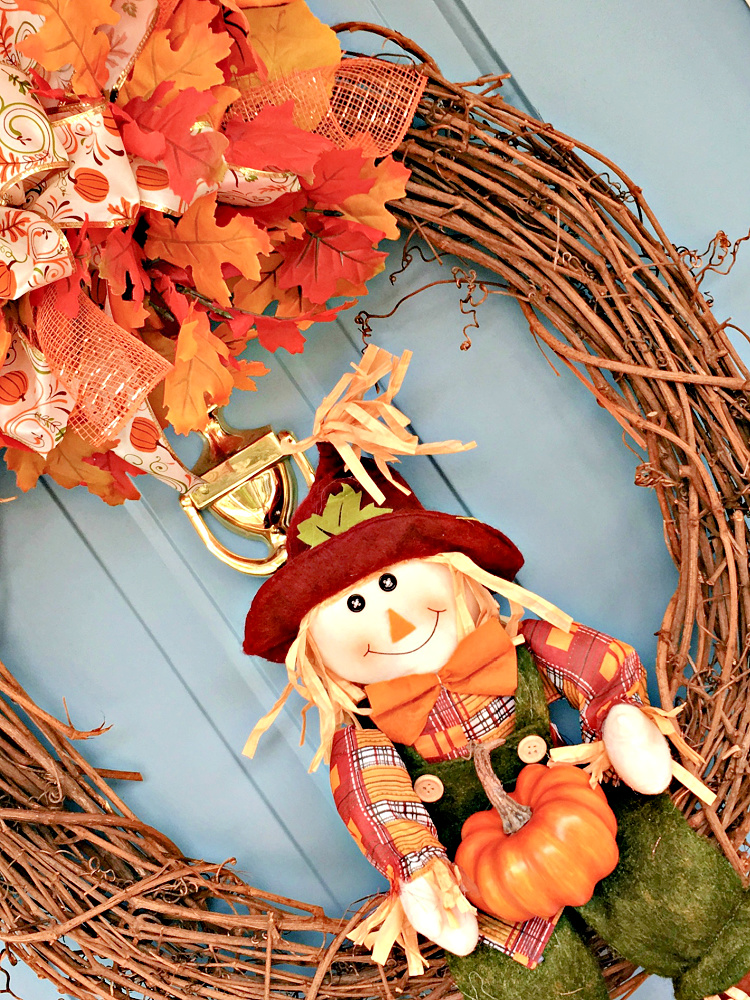 Close-up of a smiling scarecrow holding an orange pumpkin inside a fall grapevine wreath with colorful leaves, orange mesh, and pumpkin-print ribbon.