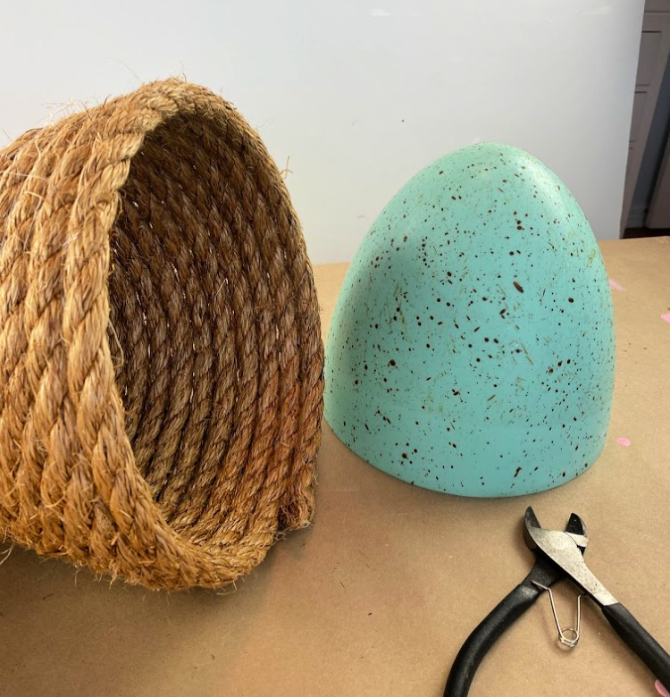 Rope bee skep on its side with half of a large blue speckled easter egg and a pair of wire cutters