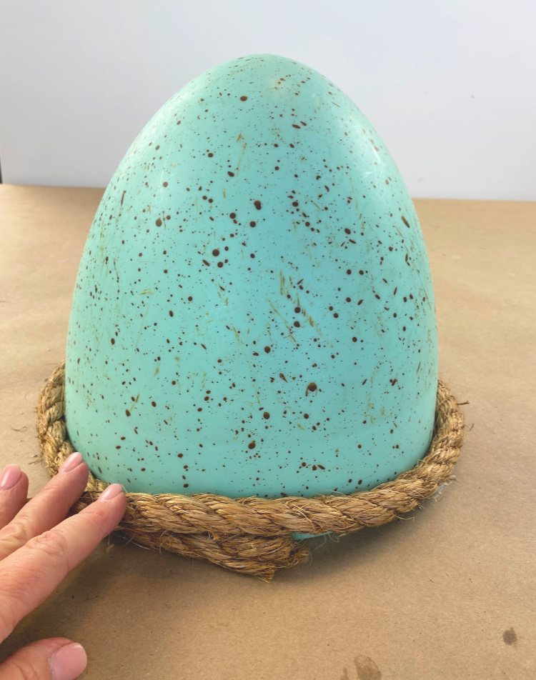 A large blue speckled easter egg with jute rope wrapped around the bottom of it. Fingers holding the rope on it