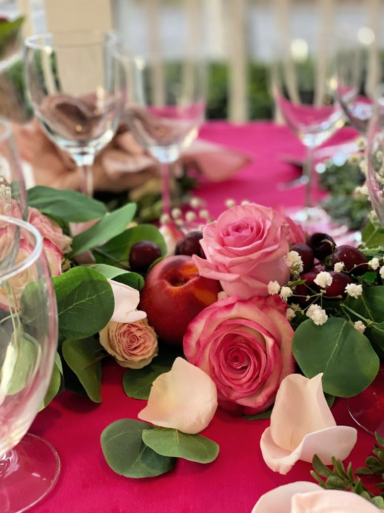 A close up photo of pink roses, babies breath, plums and cherries as a centerpiece with simple glasses blurred in the background