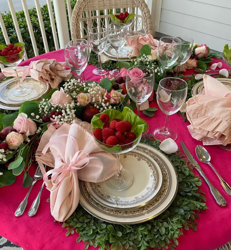 An overview of a table set with a raspberry tablecloth, 8 glass wine glasses, 4 places settings of ivory china with a delicate pattern around the edge of the china.  Silver flatware, pink napkins tied with pink ribbon.  Champagne coupes on each place setting with butter lettuce leaves and raspberries in them.  a centerpiece is running across the table with green leaves, roses in all kinds of shades of pinks and babies breath.  