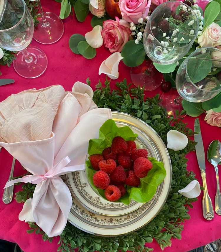 An overhead photo of a place setting with a raspberry colored tablecloth, ivory lenox china with a gray and pink floral pattern around the edge.  Silver flatware, a boxwood placemat and a bowl with raspberries resing on a bed of lettuce leaves.