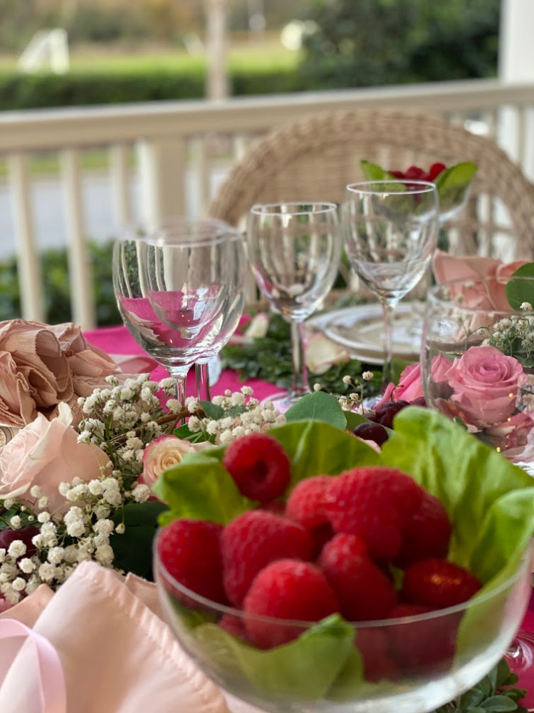 A photo of raspberries in a glass bowl on butter lettuce leaves with pink roses and baby's breath centerpiece and clear wine glasses 