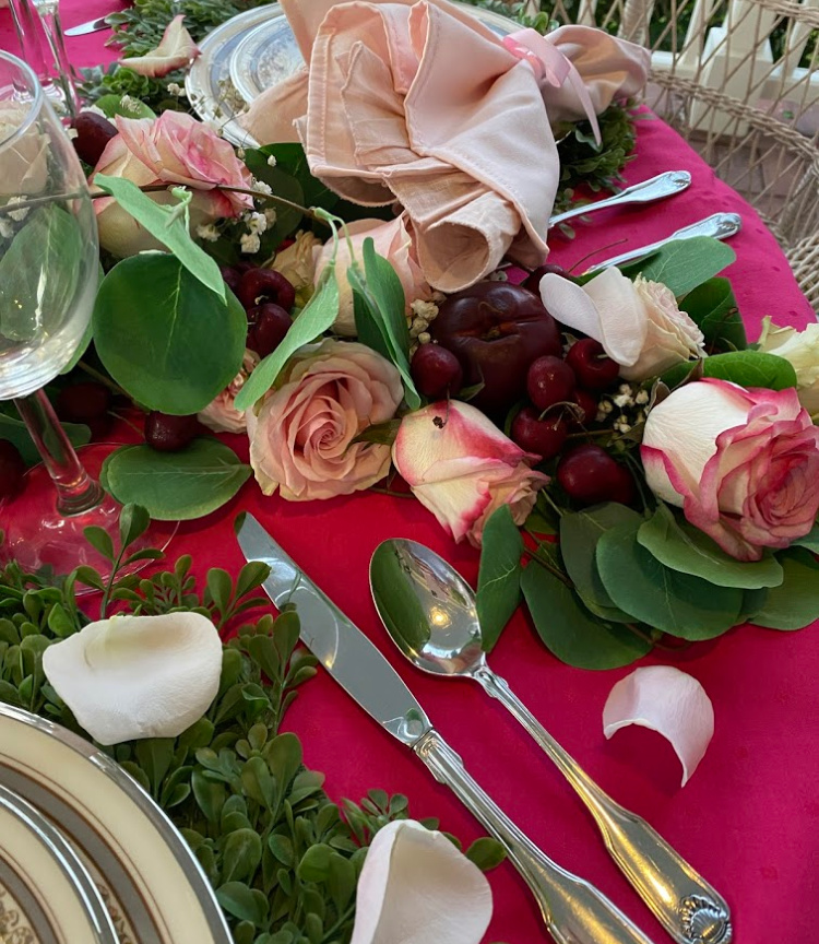An overhead shot of a ribbon centerpiece of faux eucalyptus leaves and pink roses of different shades and fresh cherries and plums. 