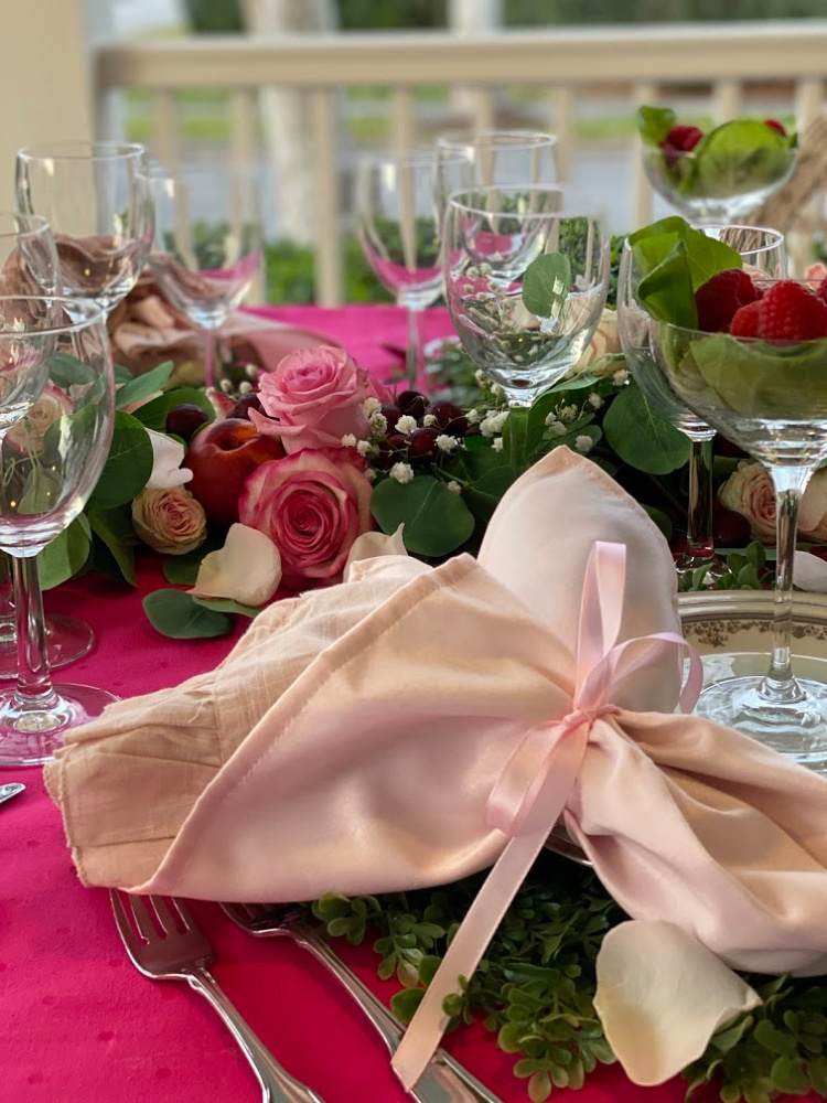 A photo of a tablescape with greenery, pink roses and pink napkins tied with a pink ribbon and several clear wine glasses.  