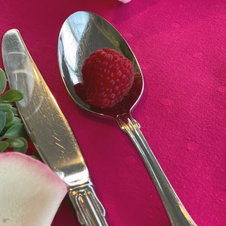 Close up of a raspberry tablecloth, a silverware knife and spoon and a raspberry sitting in the spoon.
