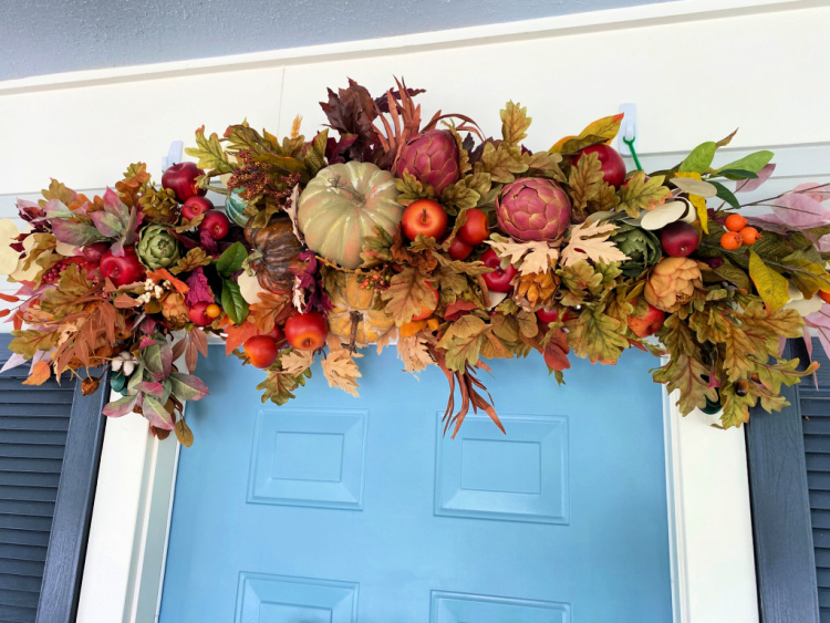 Lush fall garland filled with pumpkins, apples, artichokes, berries, and colorful autumn leaves displayed above a blue front door.