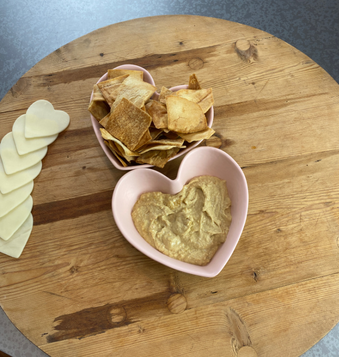 Wooden cutting board with heart shaped bowls of pita chips and hummus and cheese cut in heart shapes