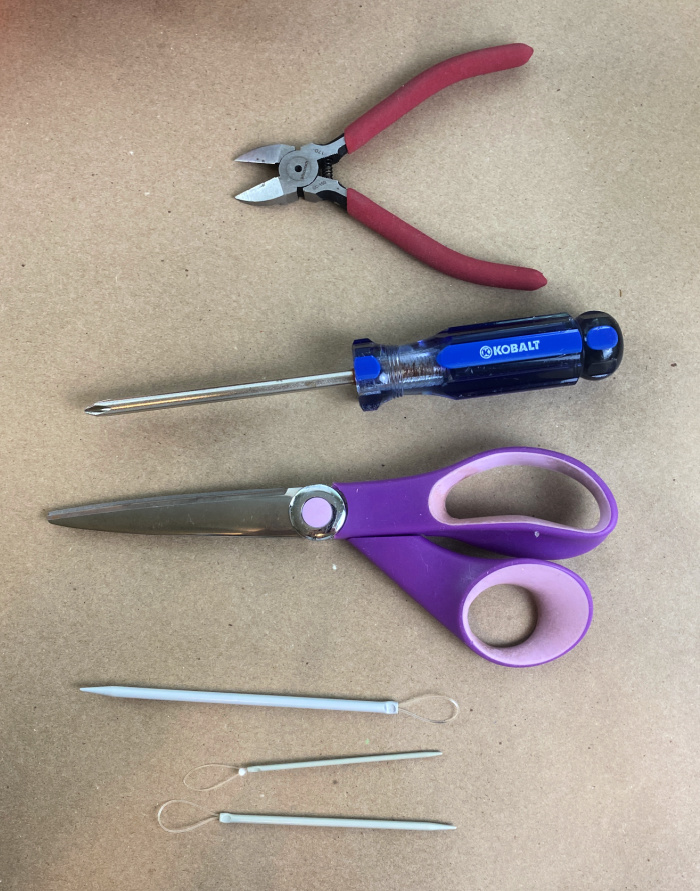 Kraft paper with a pair of purple handled scissors on it, a blue handled screwdriver, a pair of wire cutters with red handles and three flexible wool needles