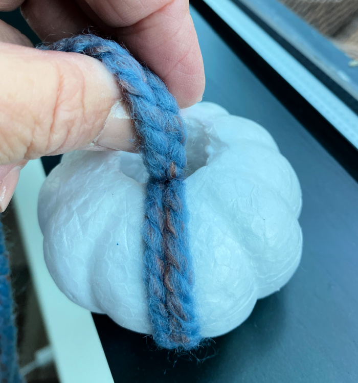 fingers holding blue yarn coming up out of the middle of a white styrofoam pumpkin with a couple of strands of blue yarn wrapped around the pumpkin