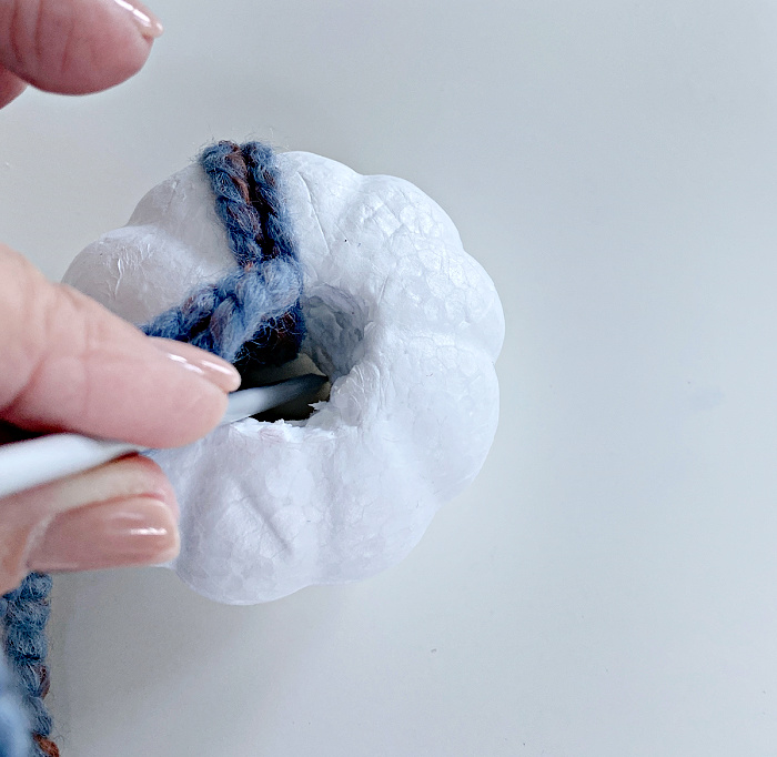 fingers holding a wool needle sticking it into a hole in the middle of a tiny white styrofoam pumpkin that has  couple of strands of blue and rust yarn wrapped around it