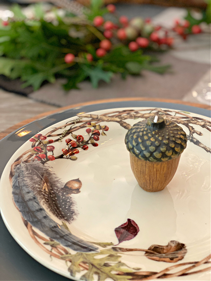 In the background, an oakleaf and berry centerpiece.  In the foreground, A place setting a faux wood charger, a gray dinner plate topped with a salad plate that is cream color with branches around the edges also decorated with berries, acorns, feathers and fungi, with a tiny acorn candle set in the middle of the salad plate.