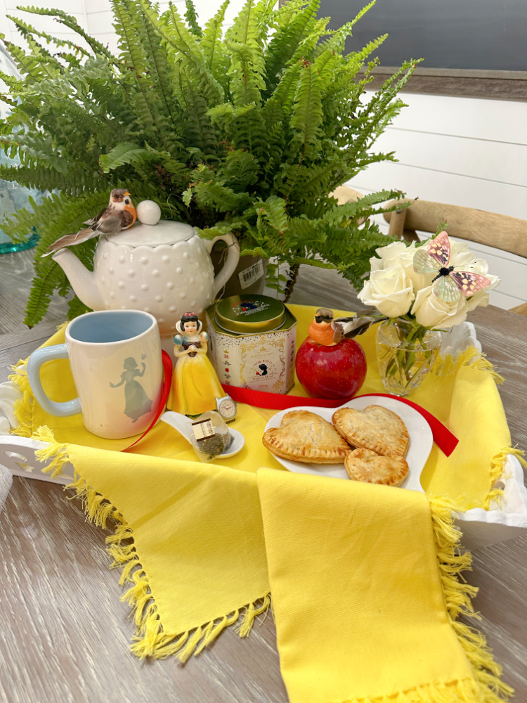 White tea tray with yellow napkins.  A green fern, a white teapot and snow white teacup with a snow white figurine and a small vase of white roses along with a white heart shaped plate with heart shaped apple pies on it. 