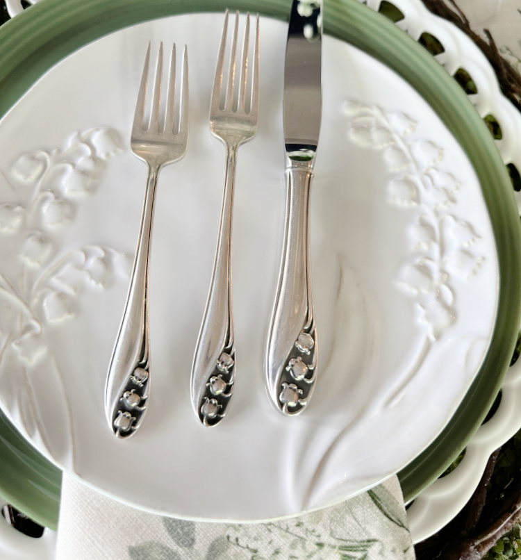 Lily of the valley silverware by Gorham, a knife, a dinner fork, a salad fork sitting across the top of a plate stack.  A white salad plate with embossed lily of the valley stems on it.  