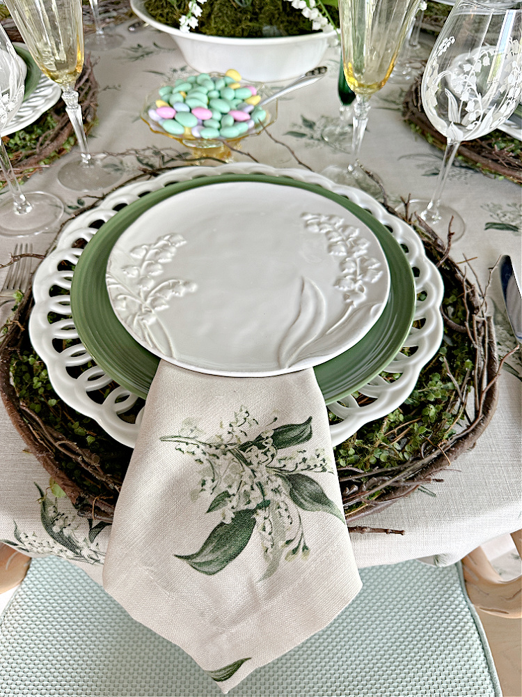 A place setting with a grapevine wreath placemat, a white openwork charger plate, a green dinner plate and a white salad plate with embossed with lily of the valley.  