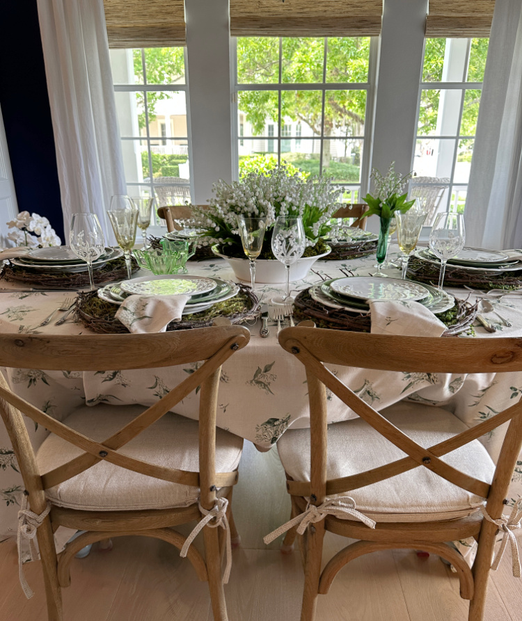 A side view of a spring tablescape showing the back of a pair of chairs and four of the place settings and a lily of the valley centerpiece.  
