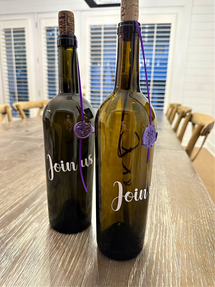 Two dark empty wine bottles with white words on them that say, "join us" with invitations rolled up inside and purple ribbons attached to the bottles with purple sealing wax.