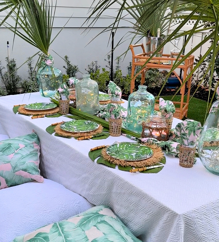 Low table in a backyard with tropical place settings and tropical and white covered pillows on the ground for people to sit upon and a rattan bar cart in the background. 