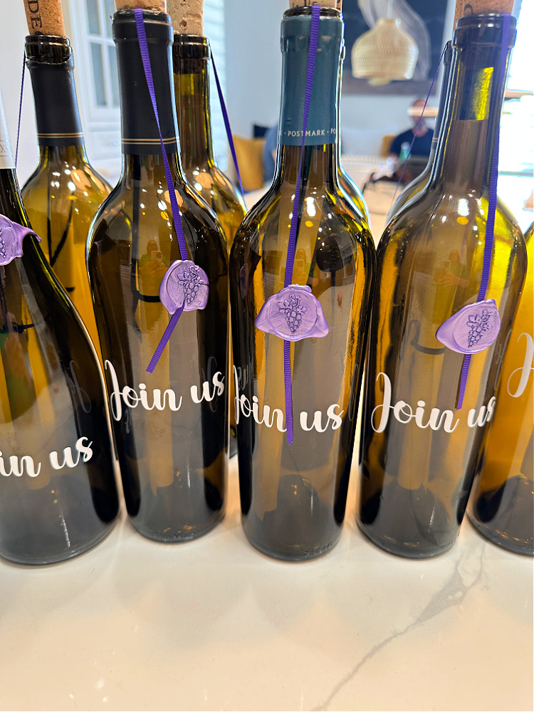 Wine bottle invitations.  Several green or brown wine bottles with invitations rolled up inside them and purple ribbons with grape sealing wax seals adhered to the wine bottles.  
