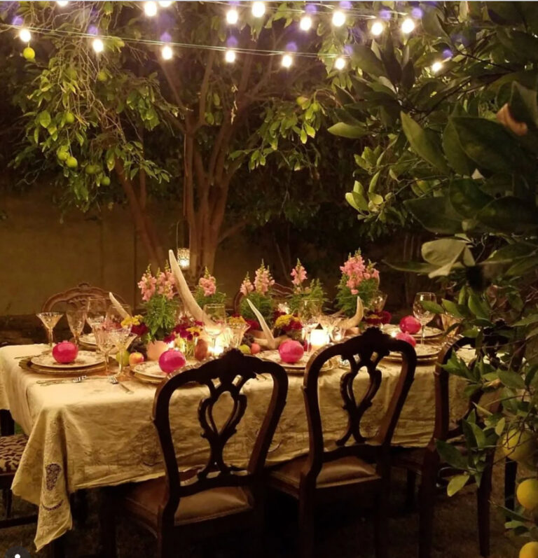 Elegant outdoor dinner table in an orchard decorated with antlers, pink flowers, fruit, vintage dishes, crystal glassware, candles, and glowing string lights.