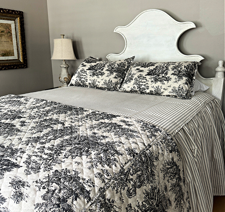 A bed with toile and striped linens on it and a sculptural pale wood headboard.  