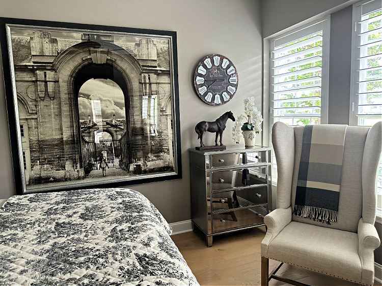 View of a French styled bedroom with a large sepia toned picture on the wall, a taupe wingback chair with a plaid throw on it.  A mirrored chest of drawers.  