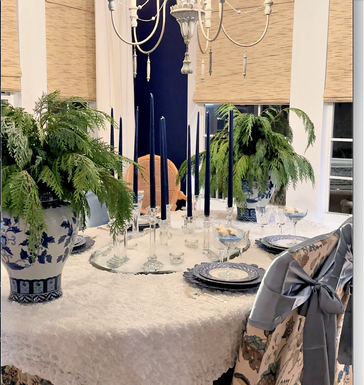 A white christmas tablescape with tall blue candles in crystal candle holders.  Slate blue bows tied on floral chairs and a pair of blue and white vases filled with cedar boughs.