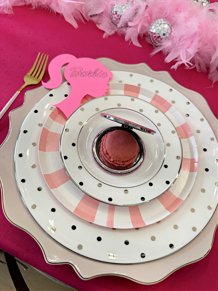 A close up photo of a Barbie themed place setting with a mirrored compact with a raspberry macaron tucked inside it.  