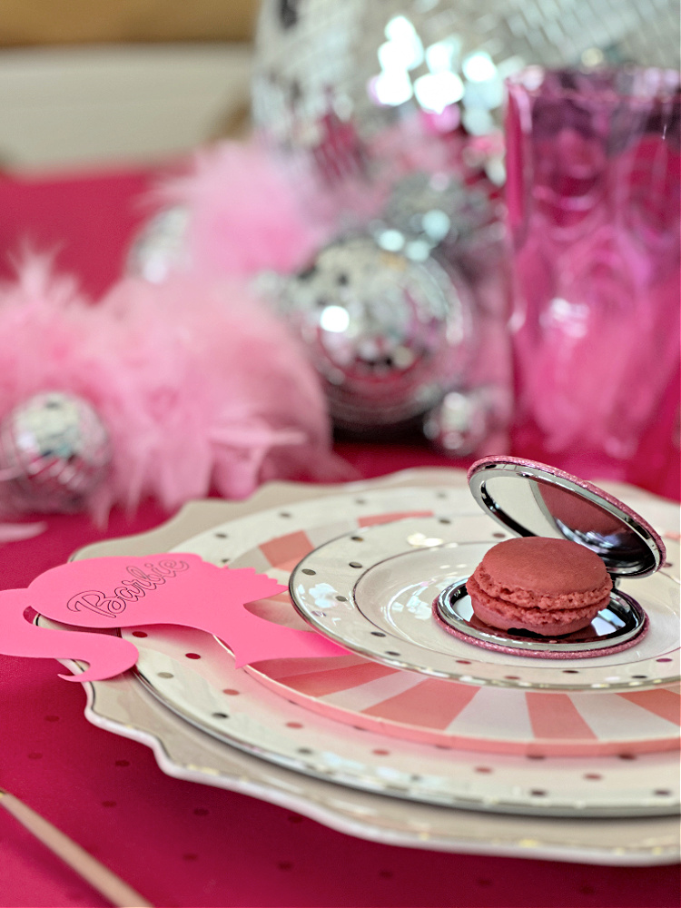 A close up photo of a Barbie themed place setting with a mirrored compact with a raspberry macaron tucked inside it.  