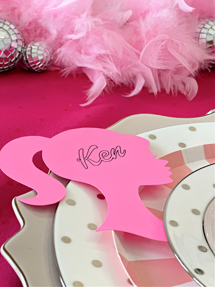 A close up of a Barbie place setting with a pink Barbie silhouette with the name Ken on it for a place card. 