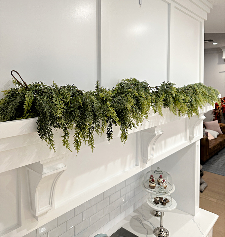 A white shelf over a cooktop in a white kitchen with a faux evergreen garland on the shelf.