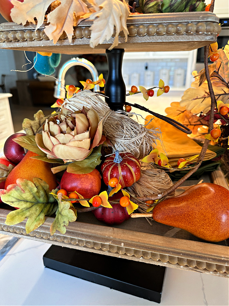 A tiered tray decorated for fall with pumpkin plates, a little plaid stuffed fox, bittersweet garland and seasonal fruits. 