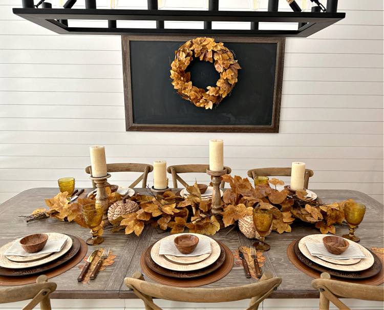 A fall table set with brown leaves, 4 candles on wooden candleholders cream colored place settings and a bulletin board with a brown leaf wreath.