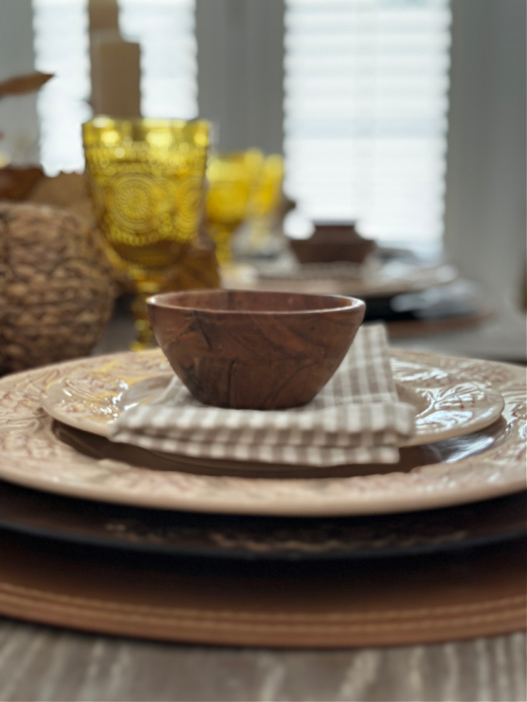 Close up of cream colored plates with a small wooden bowl sitting on them on top of a tan and white striped napkin