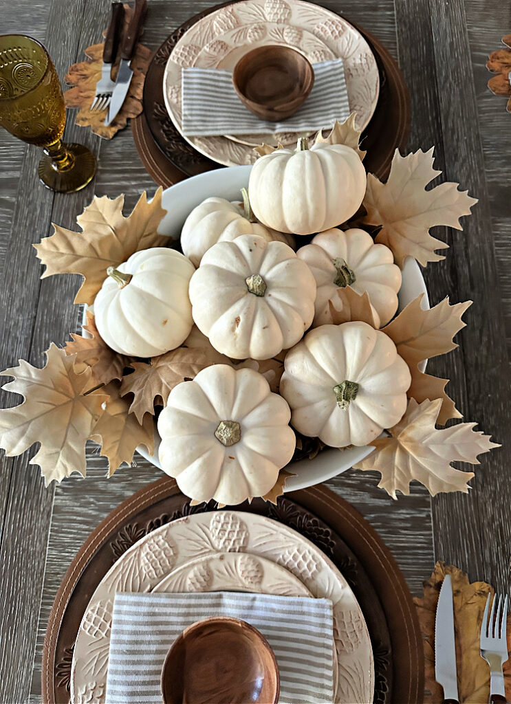 An overhead view of a white bowl with white pumpkins and pale leaves.  Two place settings with brown chargers and cream colored plates 
