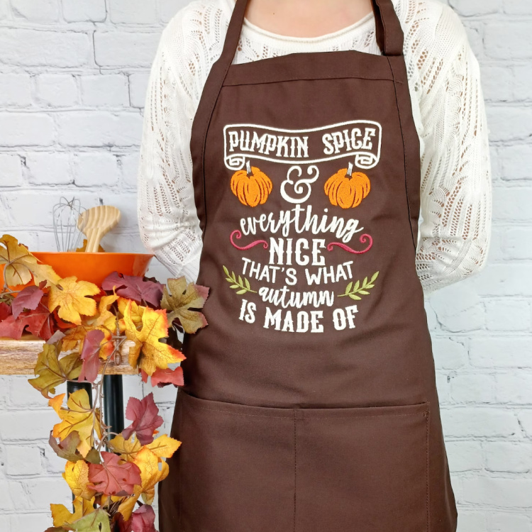 Woman's body with a white sweater and a brown apron with a pumpkin spice saying printed on it.