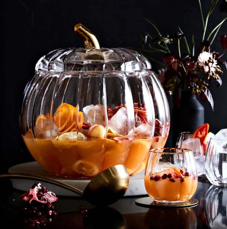 A glass pumpkin shaped punch bowl with cider punch, ice cubes and orange slices in the punch.