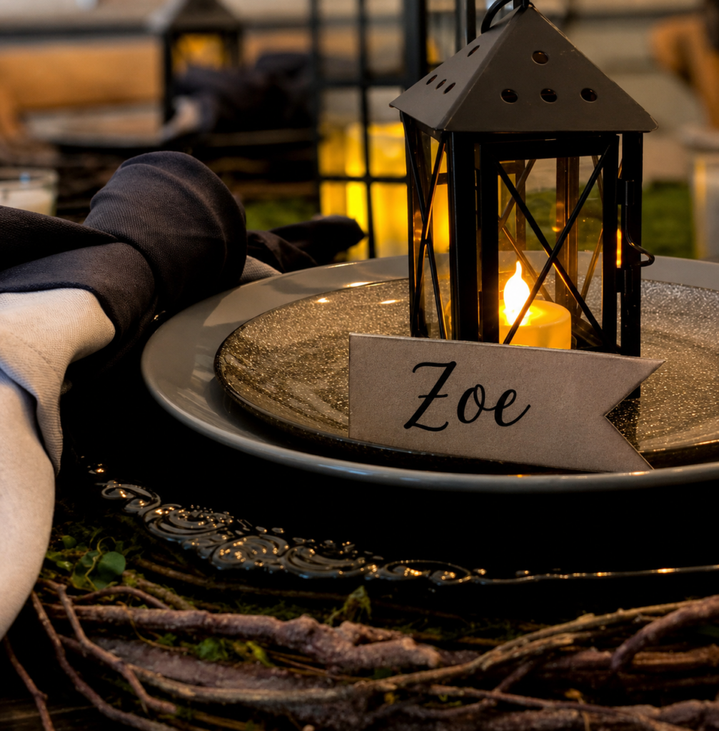 Close-up of a Halloween place setting with a black lantern, personalized place card, layered black plates, and flickering candlelight for an elegant spooky tablescape.